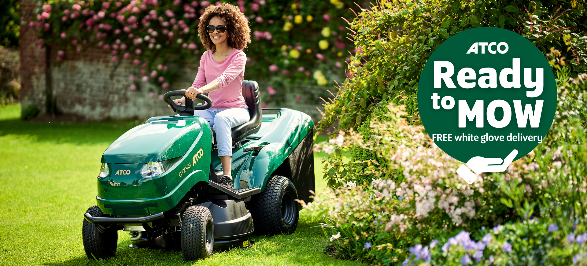 Lady riding ATCO ride-on mower in garden with 'ready to mow' logo banner